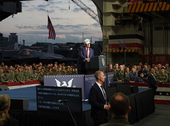 Trump, vestindo um terno escuro, gravata vermelha e boné branco que diz “EUA”, está em um estrado. Em dois lados, são vistas multidões de pessoas com roupas militares camufladas.