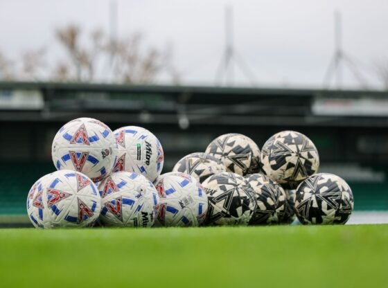 Uma pilha de bolas de futebol brancas, vermelhas e azuis e uma pilha de bolas de futebol pretas e brancas num campo verde.