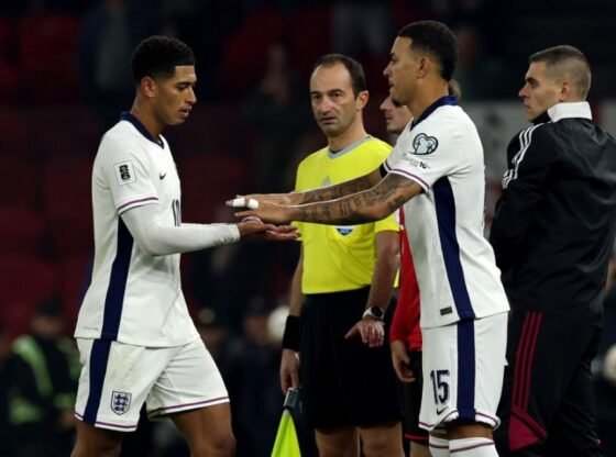 Dois jogadores de futebol da Inglaterra com camisas brancas se enfrentam em campo com um árbitro de camisa amarela atrás deles.
