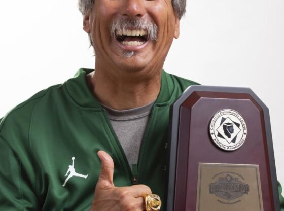 O técnico do Laney College, John Beam, sorri e gesticula com a mão enquanto segura o troféu do campeonato estadual.